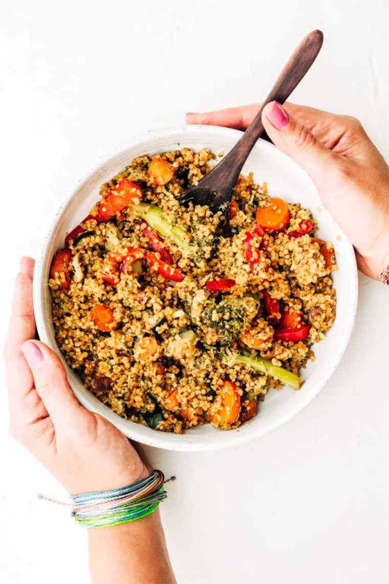 two hands holding a white bowl with oil-free stir fried quinoa, carrot, bell pepper and broccoli