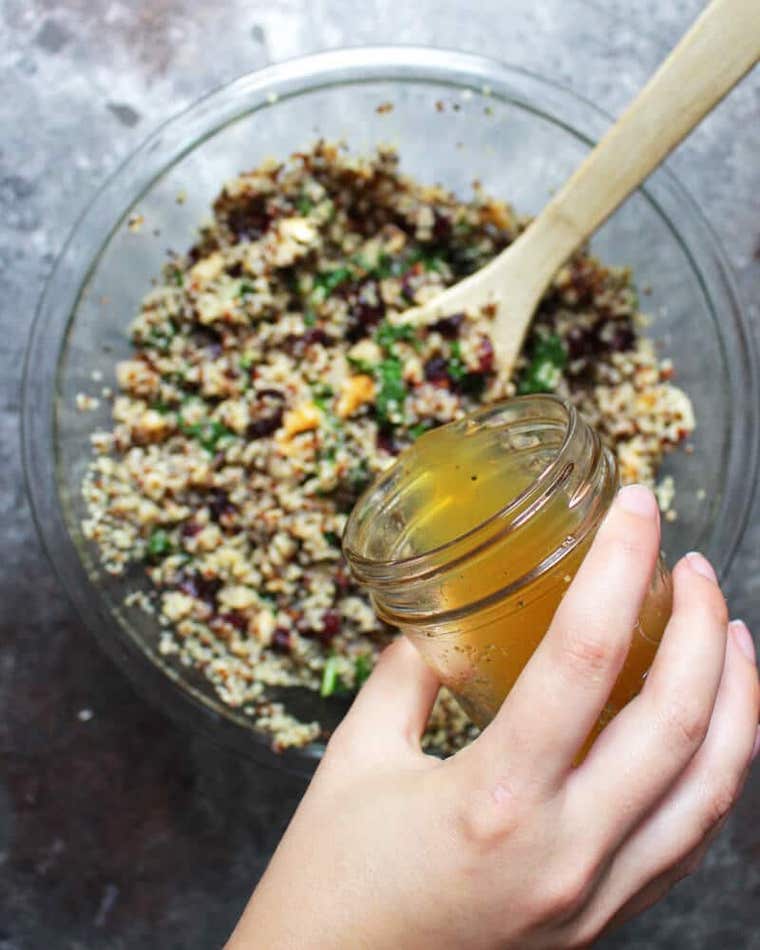 large glass bowl with a colorful plant based grain salad in which some light apple sage dressing is being poured by a hand