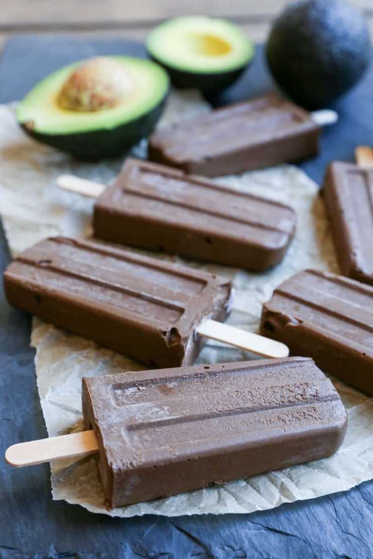 dark table with some parchment paper and avocados next to five raw vegan chocolate fudgesicles