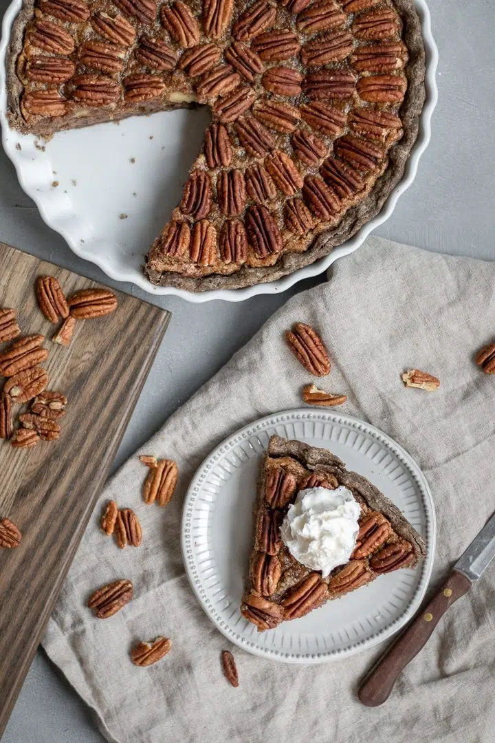 white pie form with a whole food plant based vegan pecan pie of which a piece has been cut out and placed on a small plate next to it, topped with vegan whipped yream