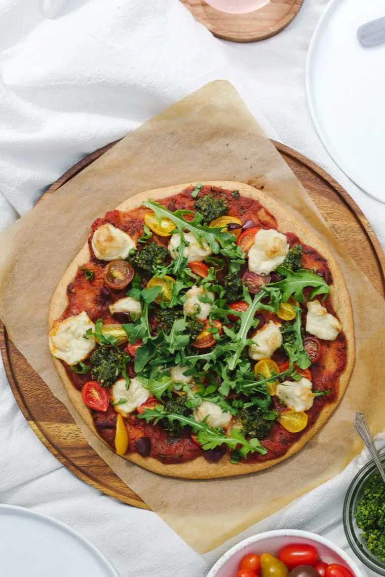 round wooden chopping board with parchment paper and a vegan quinoa pizza crust loaded with tomatoes and arugula