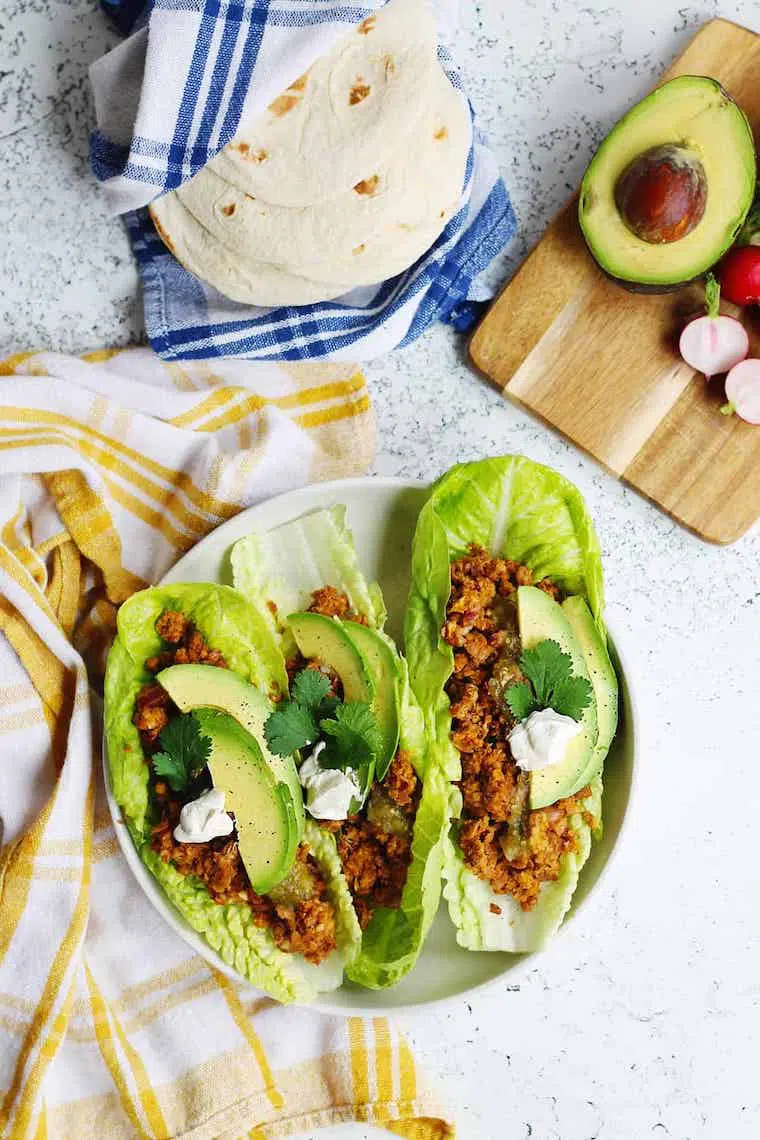 white table with two towels, a chopping board and a white plate with vegan taco lettuce wraps and avocado