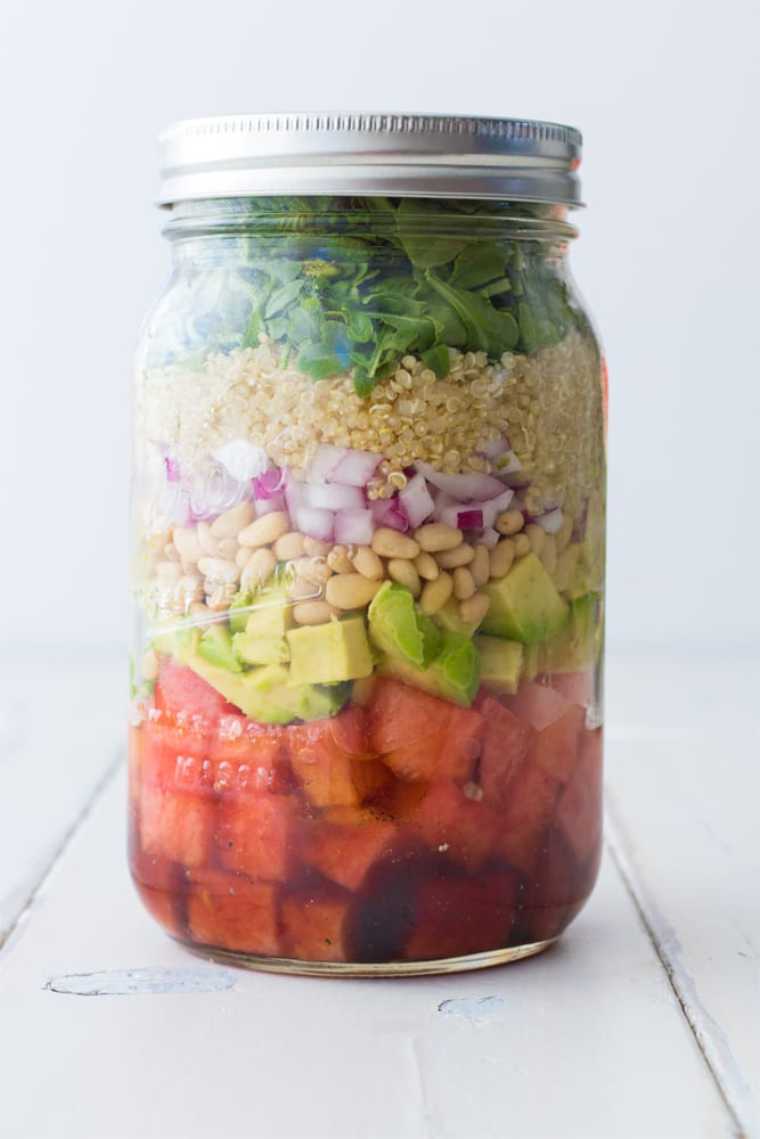 white table with a glass mason jar containing cubed watermelon, avocado, white beans, red onion, quinoa and leafy greens