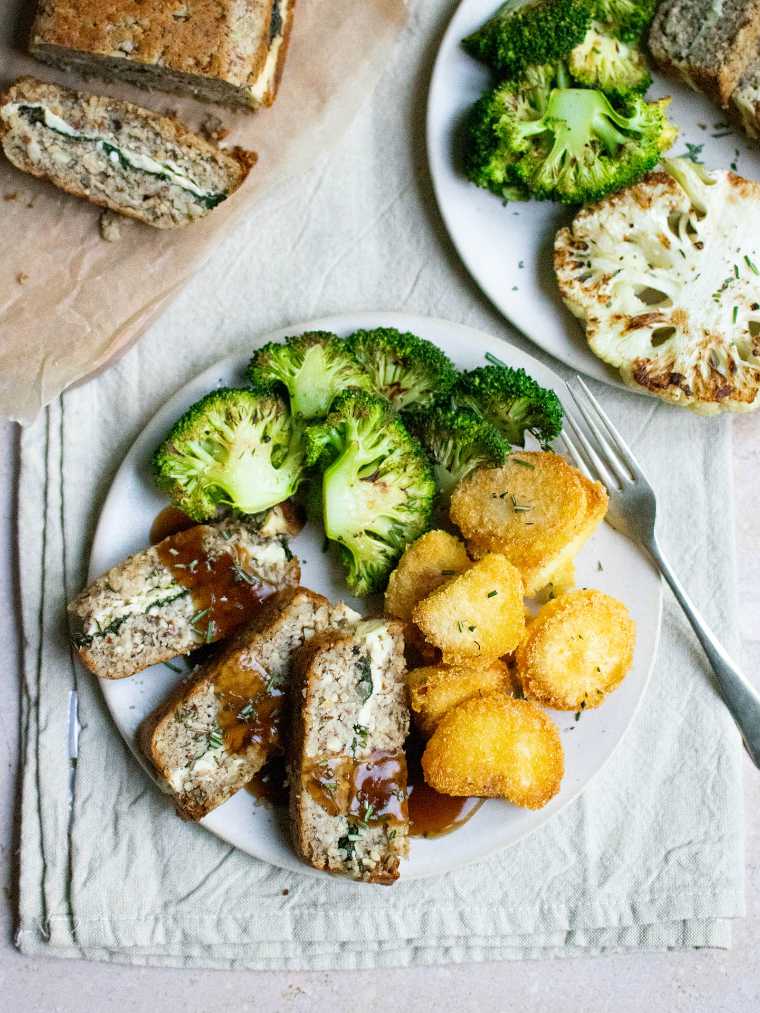 two white plates with roasted broccoli, potatoes and vegan nut roast