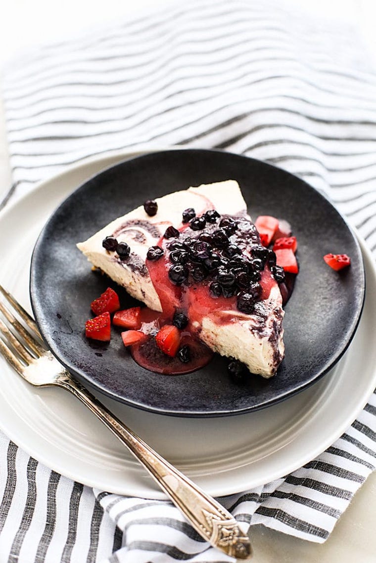 black plate on a white table with a piece of no-bake vegan cheesecake with a berry swirl and topped with strawberries and blueberries