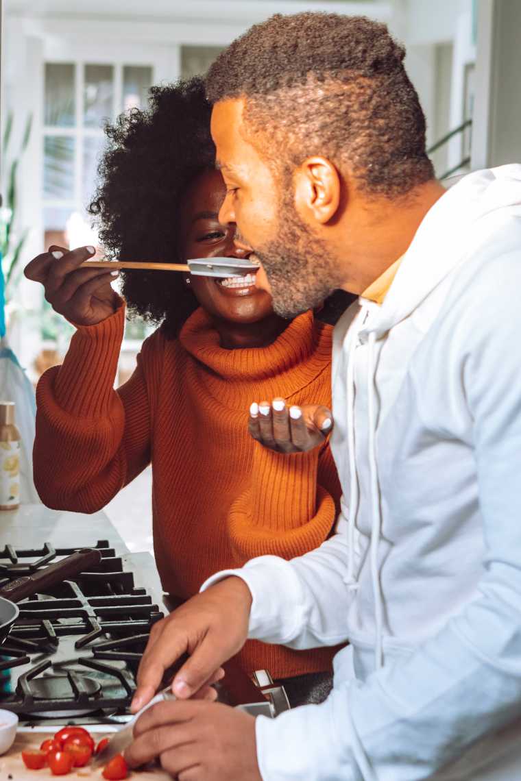 African American woman and man in kitchen with man cutting tomatoes on a cutting board and woman holding spatula near man's mouth to let him try some food