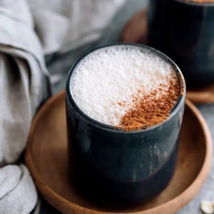two cups of frothy vegan latte topped with cinnamon next to a linen towel