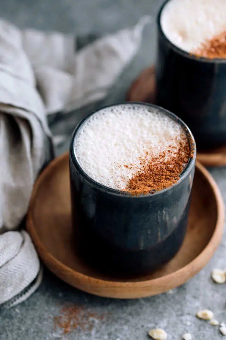 two cups of frothy vegan latte topped with cinnamon next to a linen towel