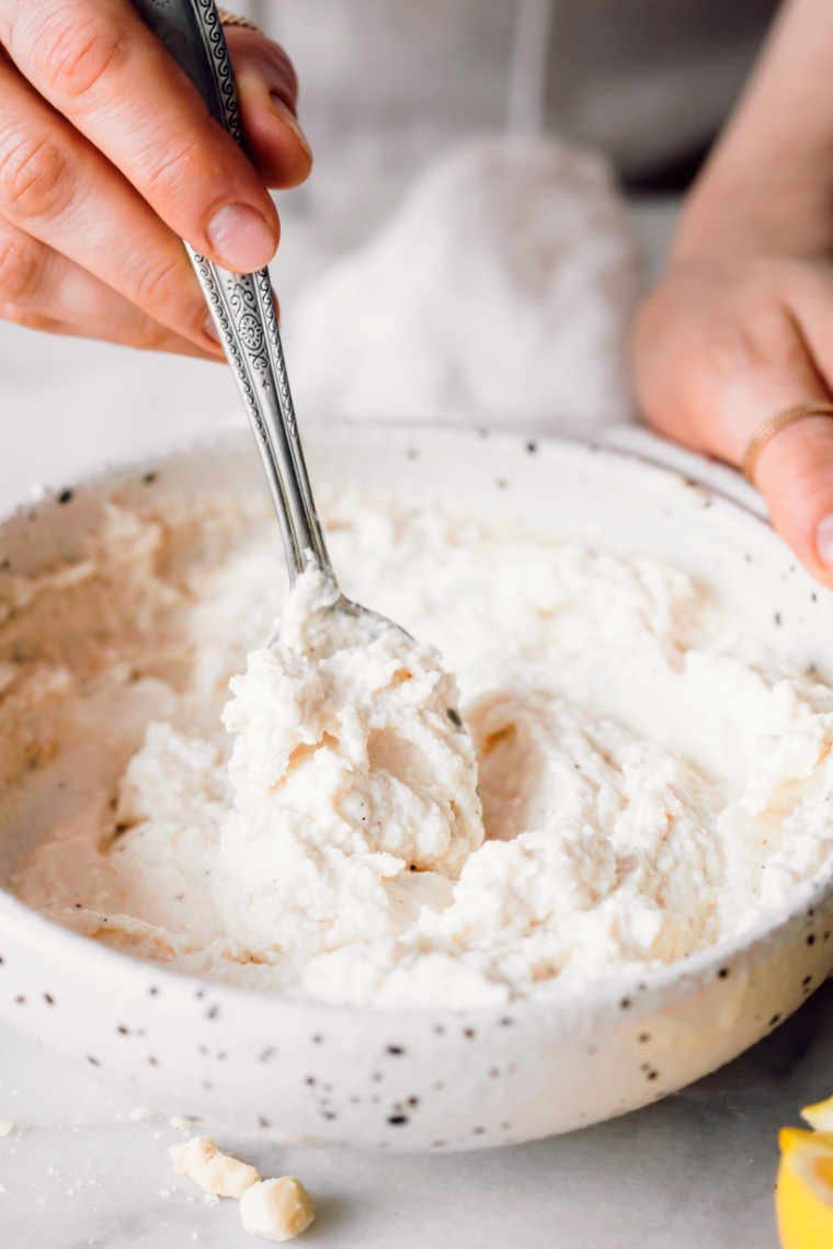 woman holding white bowl with almond ricotta in her hand and a spoon in the other with which she mixes the ricotta