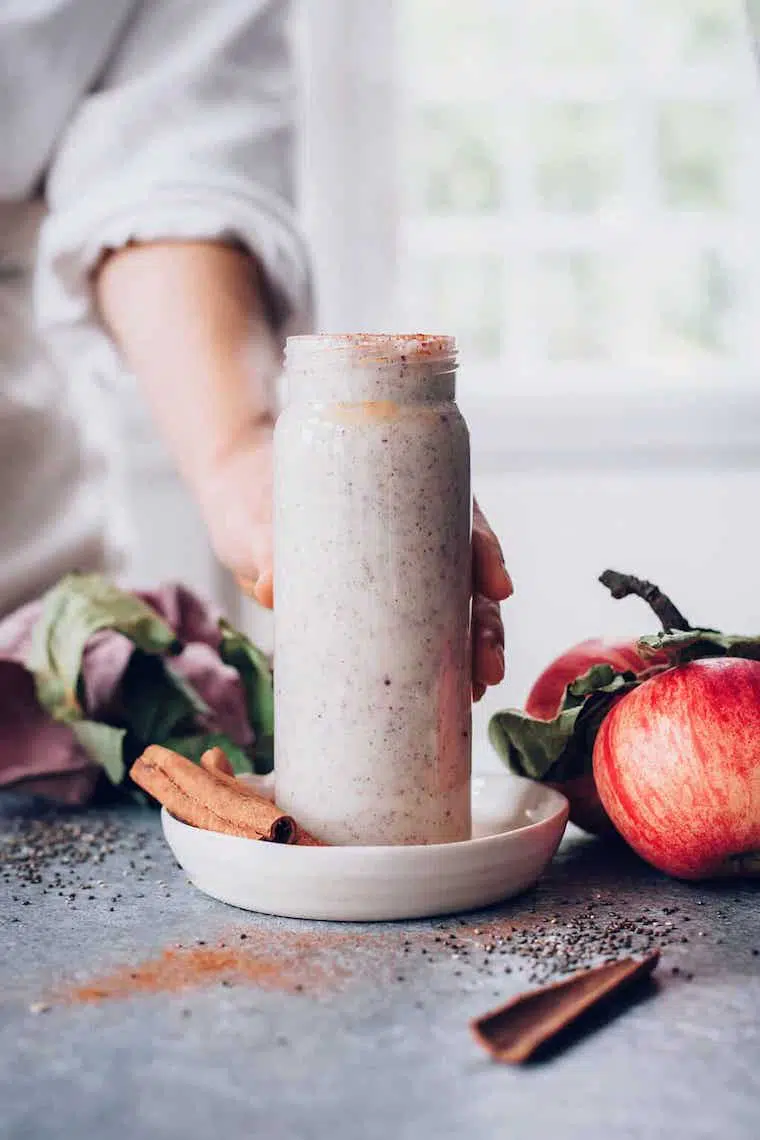 woman in white shirt holding a glass with purple applie pie chia smoothie next to apples and cinnamon