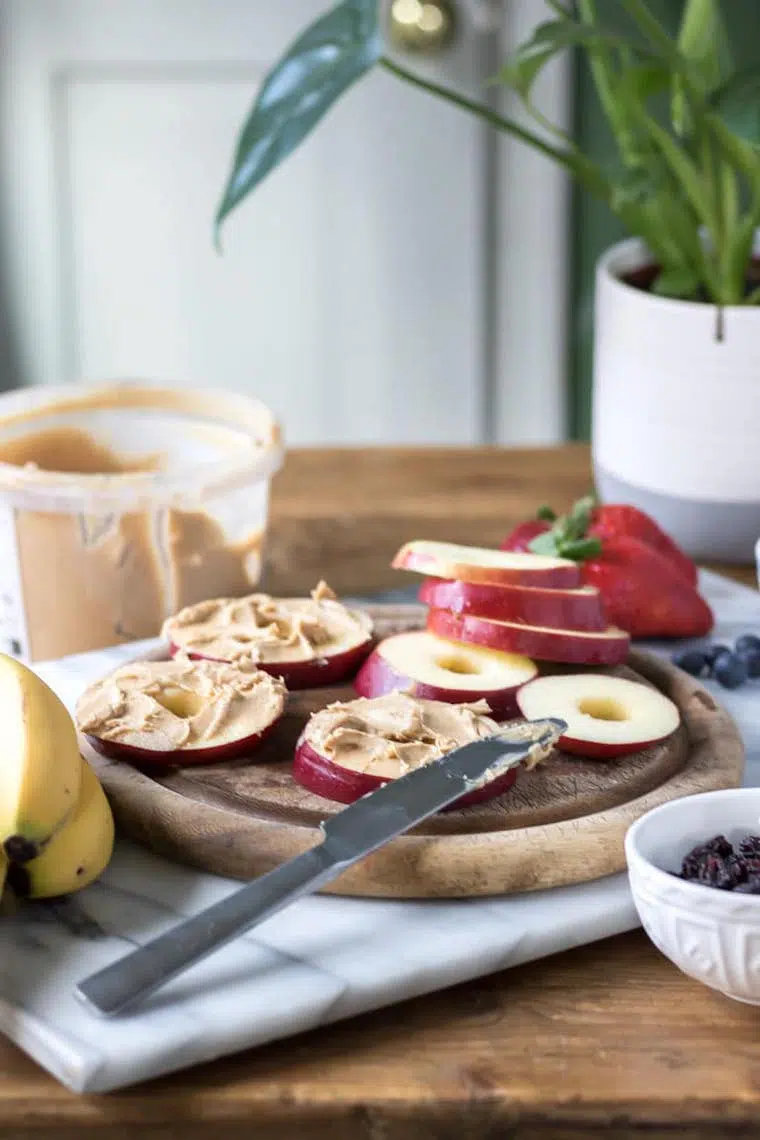wooden table with a jar of peanut butter and a chopping board with apple slices that are being spread with peanut butter