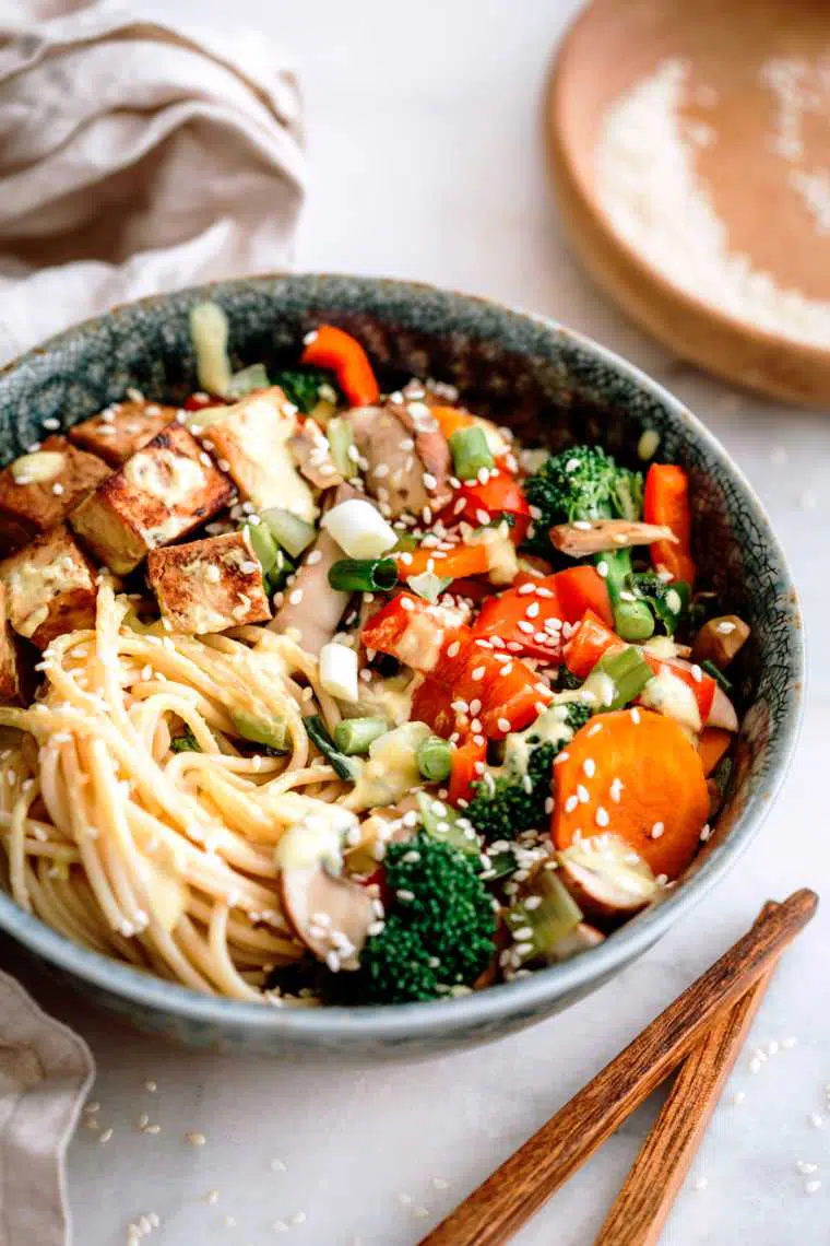 close up of a large bowl with cooked noodles and different Asian veggies, such as carrot, bell pepper and broccoli as well as some marinated tofu next to two chopsticks