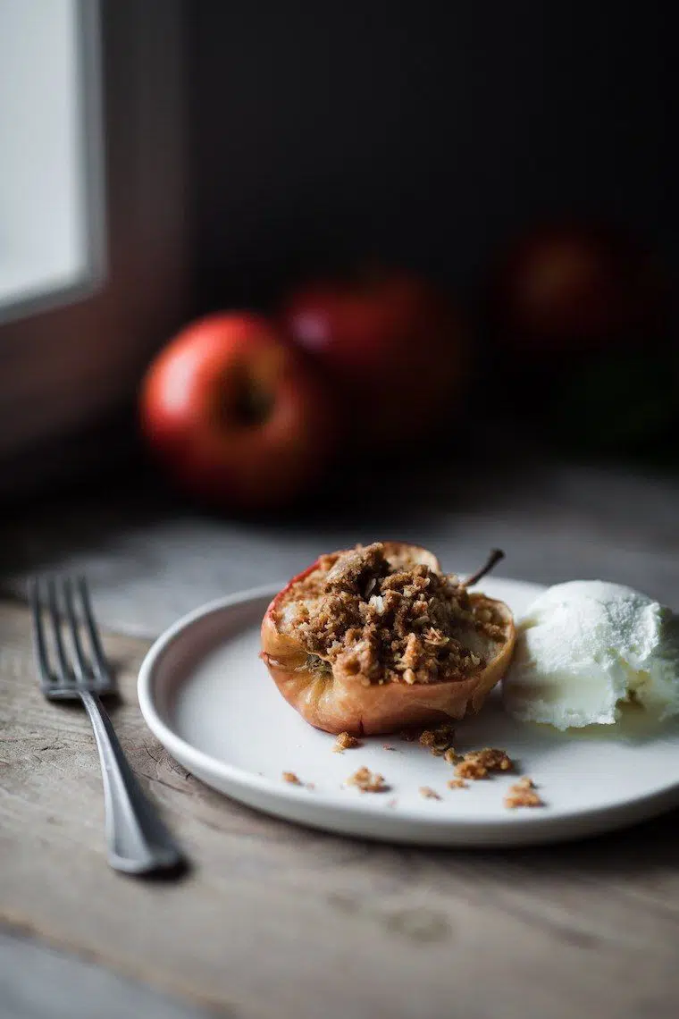 white plate on wooden surface with half a baked apple stuffed with oat streusel