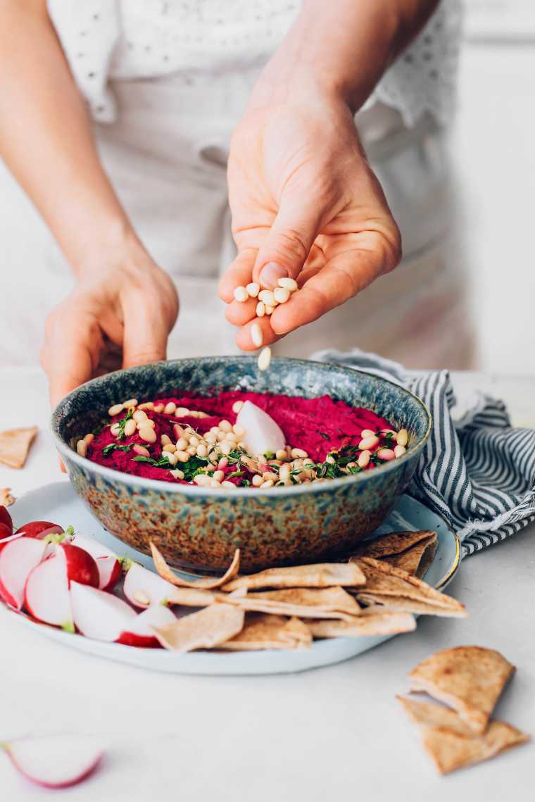 woman standing by a table and topping beetroot dip with pine nuts
