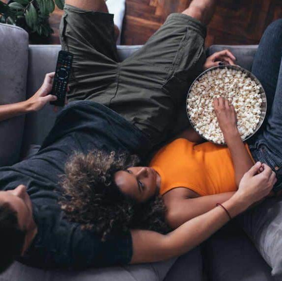 Overhead view of a couple on a grey couch. Woman eating popcorn and lying down in a man's arm who is holding a remote control in his other hand.