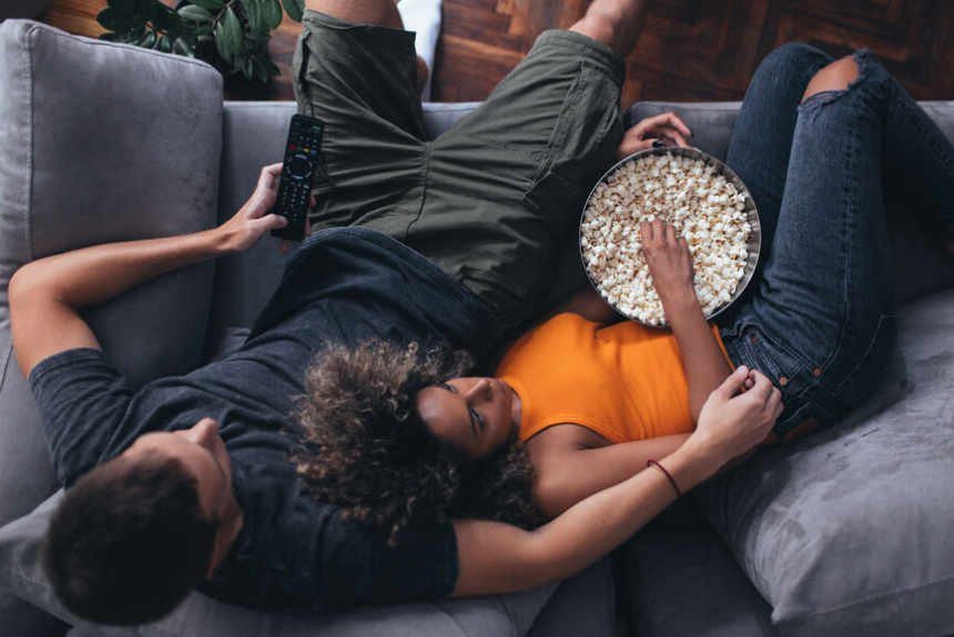 Overhead view of a couple on a grey couch. Woman eating popcorn and lying down in a man's arm who is holding a remote control in his other hand.
