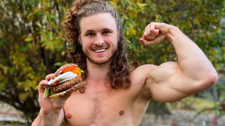 Brian Turner smiling and showing off his right biceps while holding vegan sandwich with his left hand