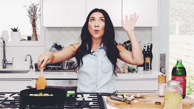 Lauren from Hot For Food standing in front of stove with funny expression, throwing her left arm into the air while stirring vegetables in a pan with her right arm