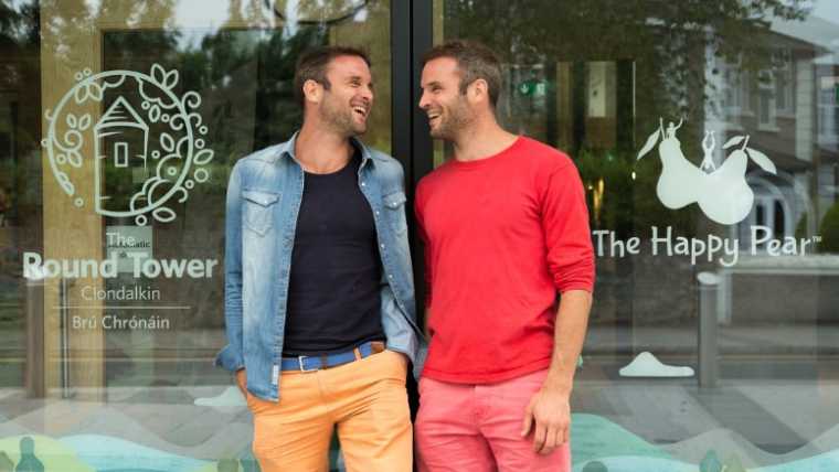 Dave and Steve from The Happy Pear in colorful clothes standing in front of shop entrance, looking at each other and laughing