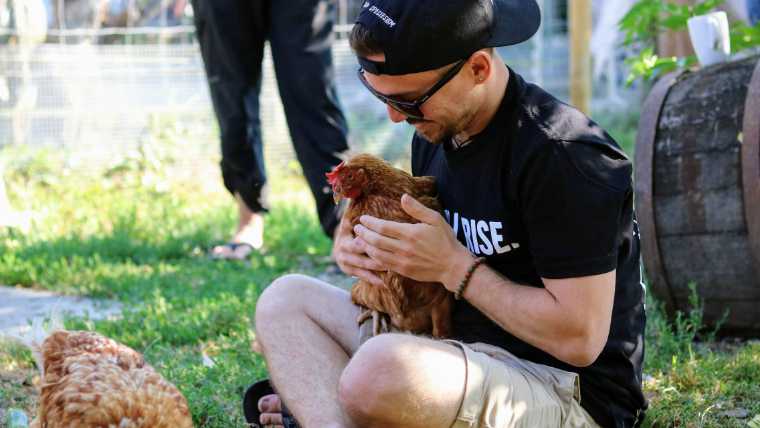 Joshua Entis sitting cross-legged on a lawn, holding a chicken in his hands