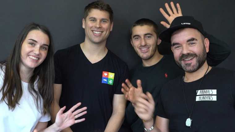 The team of Plant Based News standing in front of black wall, smiling and waving into the camera