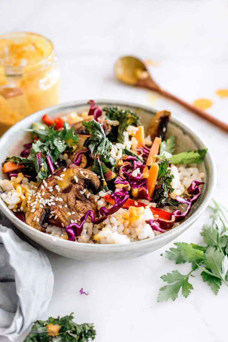 close up of a white bowl containing rice, mushrooms, red cabbage, sliced carrot, bell pepper and kale next to a creamy miso dressing