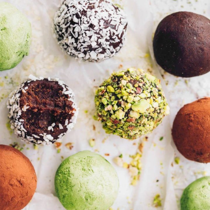 different colored bliss balls with matcha, coconut, cocoa and pistachios on white parchment paper