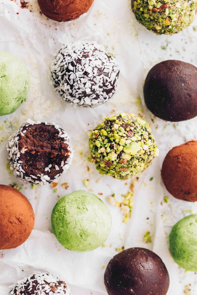 different colored bliss balls with matcha, coconut, cocoa and pistachios on white parchment paper