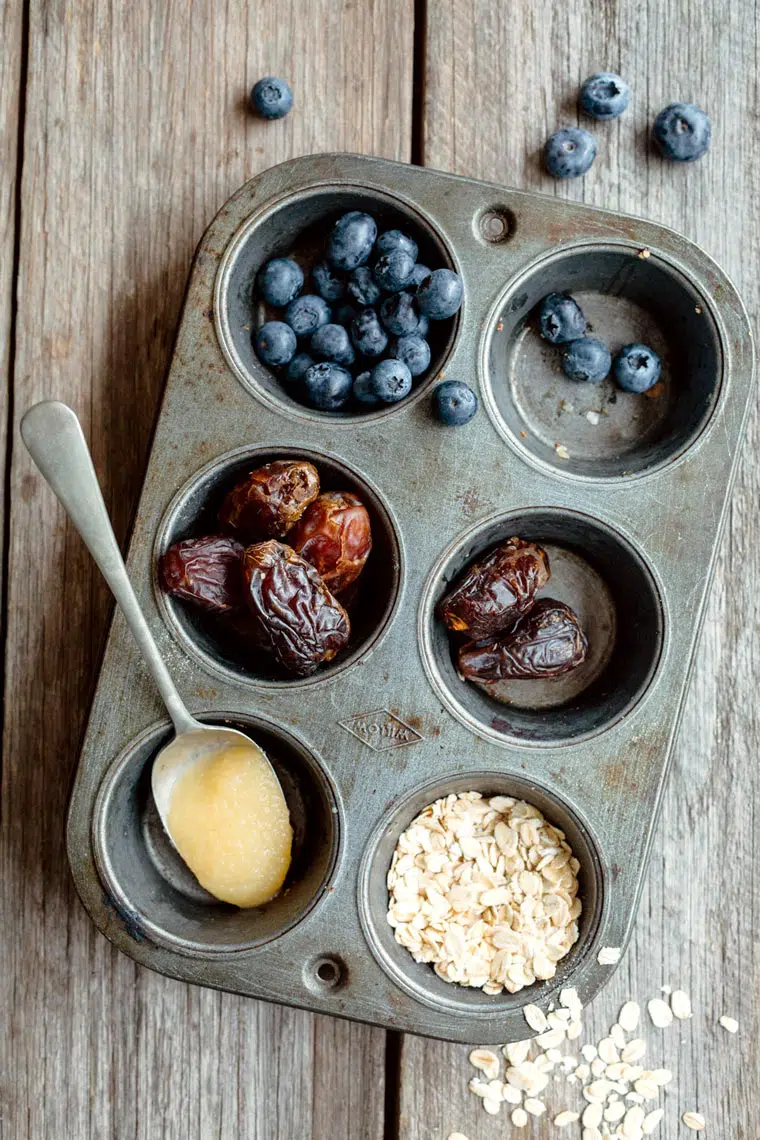 top view of a muffin tin with different ingredients that are used to make gluten-free blueberry muffins, such as dates, applesauce and oats