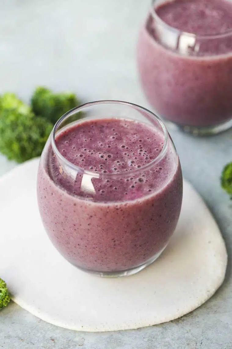 two glasses of purple blueberry broccoli smoothie next to some broccoli florets
