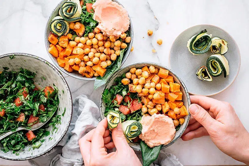 two hands placing zucchini rolls in a bowl with sweet potato, chickpeas, greens and hummus