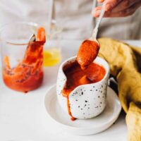 woman in apron standing by a table and pouring homemade Buffalo sauce from a spoon into a jar