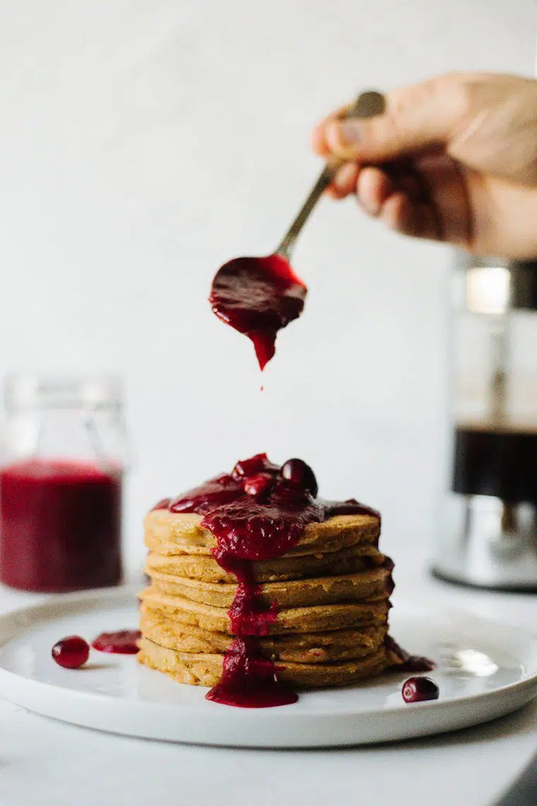 stack of vegan cardamom pancakes with a hand putting cranberry compote on top