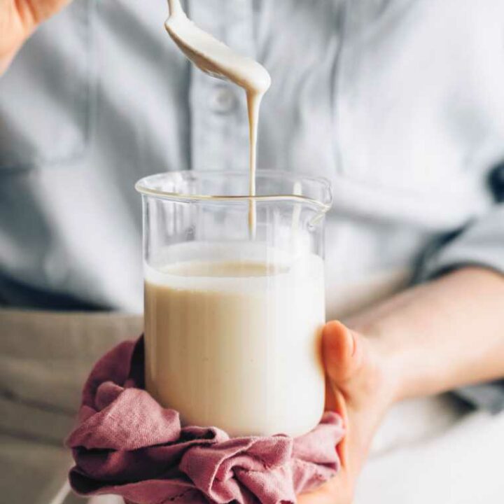 woman in blue shirt and apron holding a glass jar with homemade cashew sour cream which is dripping from a spoon