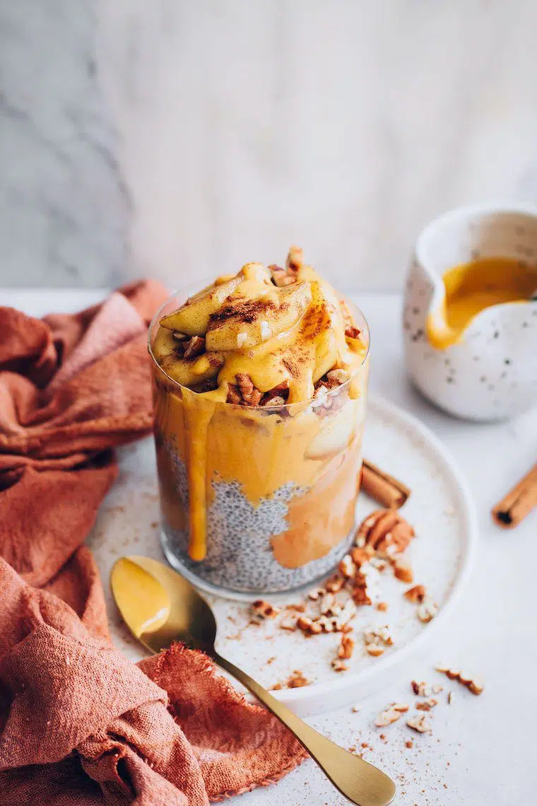 glass of chia pudding with yellow pumpkin caramel and topped with nuts on a white plate next to a spoon