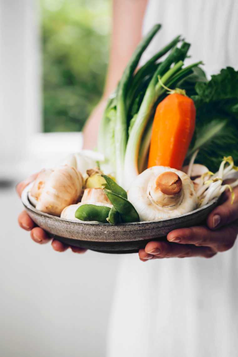 woman in a white dress holding a bowl with vegetables like mushrooms, carrots, Bok Choy and scallions