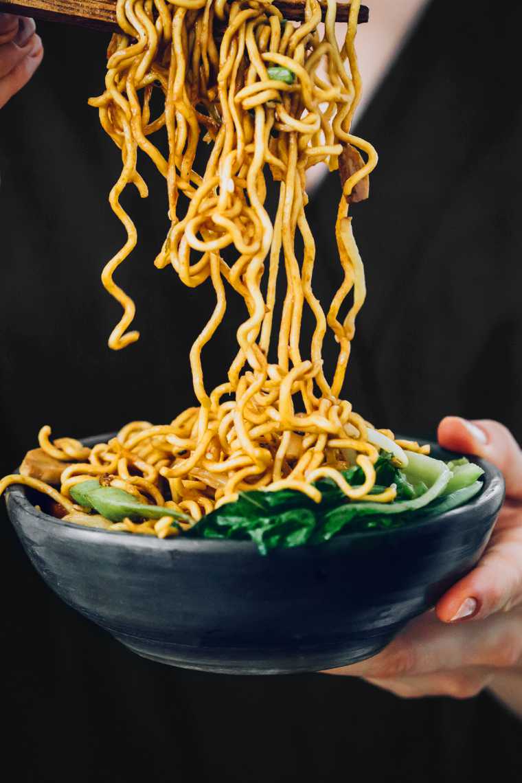 woman in dark clothes holding a bowl of homemade vegan Chow Mein noodles and eating them