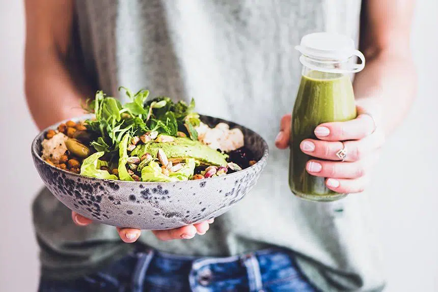 woman in green shirt and jeans holding a grey speckled bowl of green vegetables and beans in one hand and a green smoothie in the other
