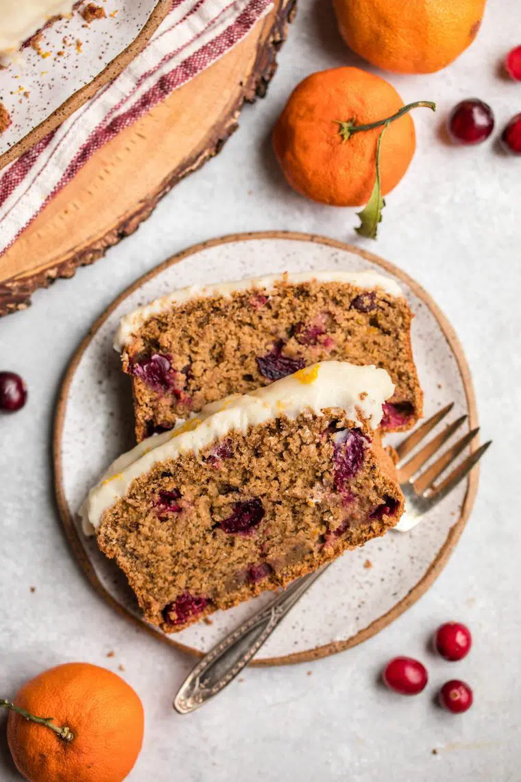 two slices of moist cranberry orange bread with white glaze on a white plate next to oranges and a fork