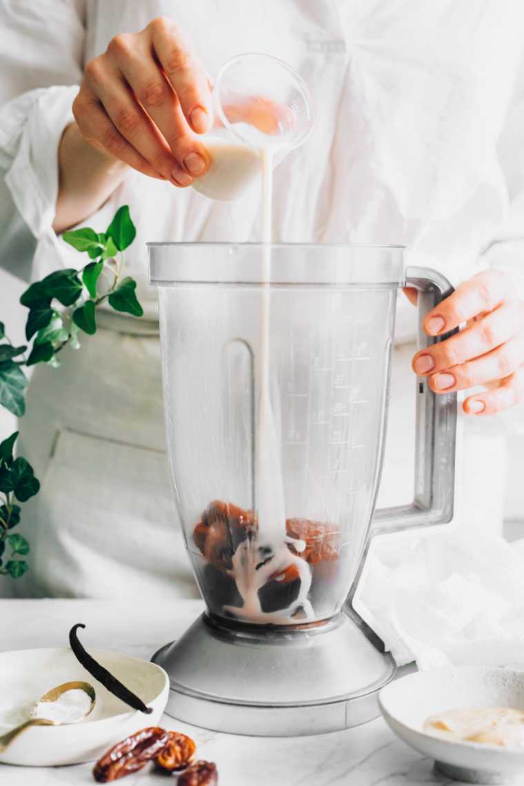 woman in white shirt and apron standing next to a blender and pouring almond milk over dates to make vegan caramel