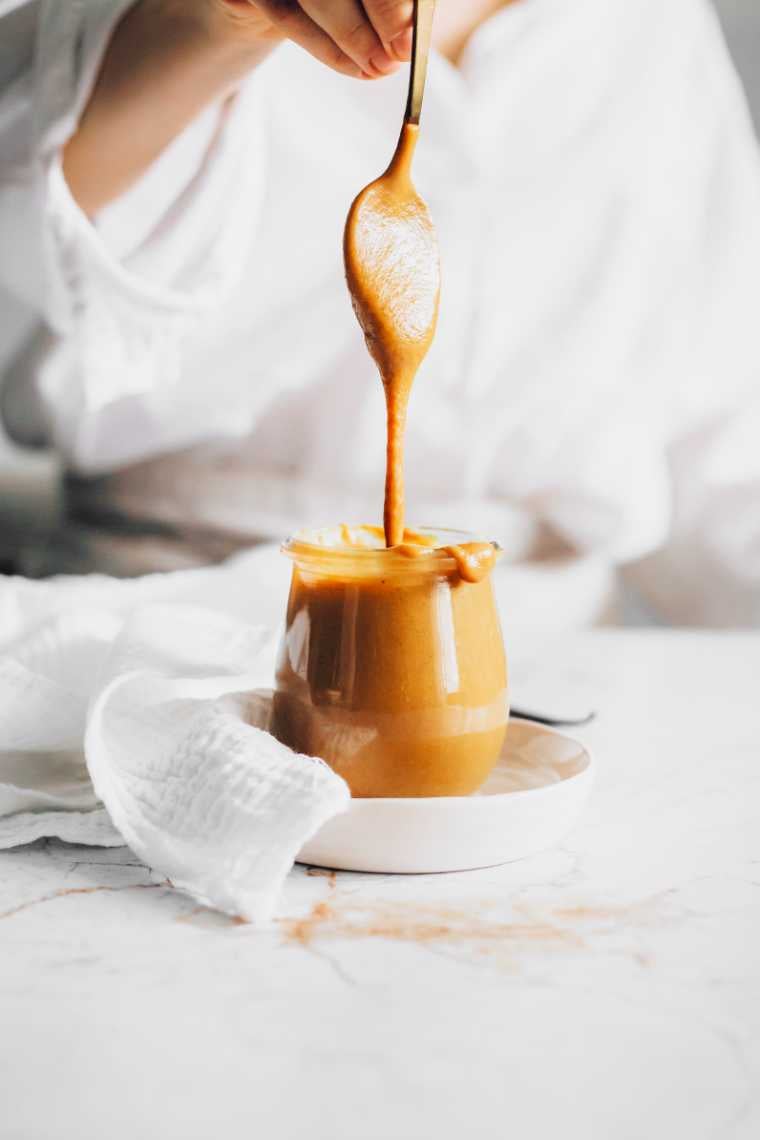 woman in white shirt standing next to a white table with a glass jar of homemade vegan date caramel with a spoon