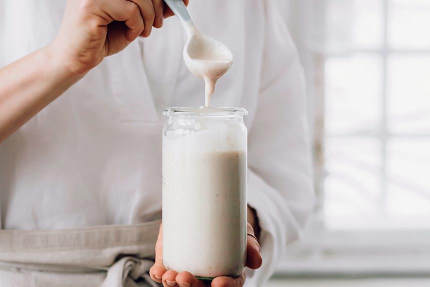 woman in kitchen holding a glass of white vegan cheese sauce with a white spoon in her hand