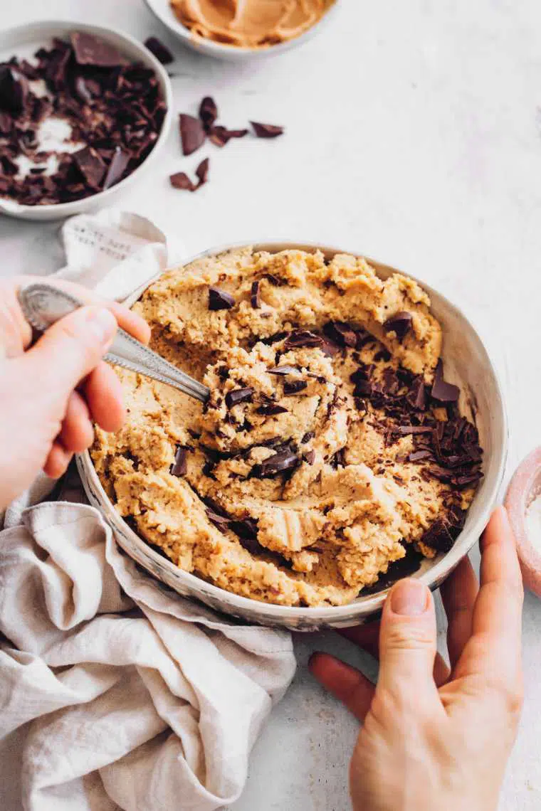 bowl on a table in which chopped chocolate is being mixed into protein cookie dough with a spoon