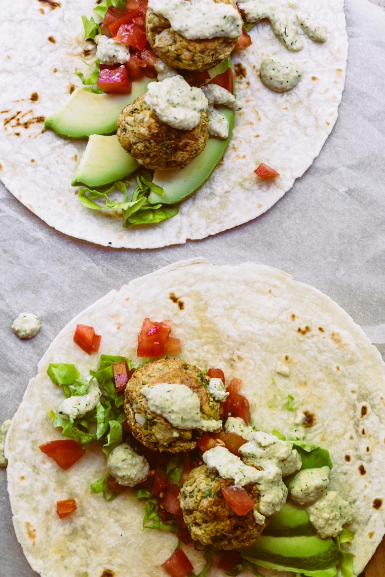 two tortilla on parchment paper that are being filled with leafy greens, tomatoes, oil free baked falafel and cashew sour cream