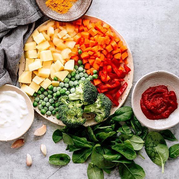 plate with potato, carrot, bell pepper, broccoli and peas standing next to small bowls of soy yogurt, spices, tomato paste and fresh spinach