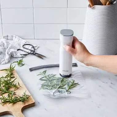 marble kitchen counter on which a hand uses a small tool to vacuum seal some fresh herbs