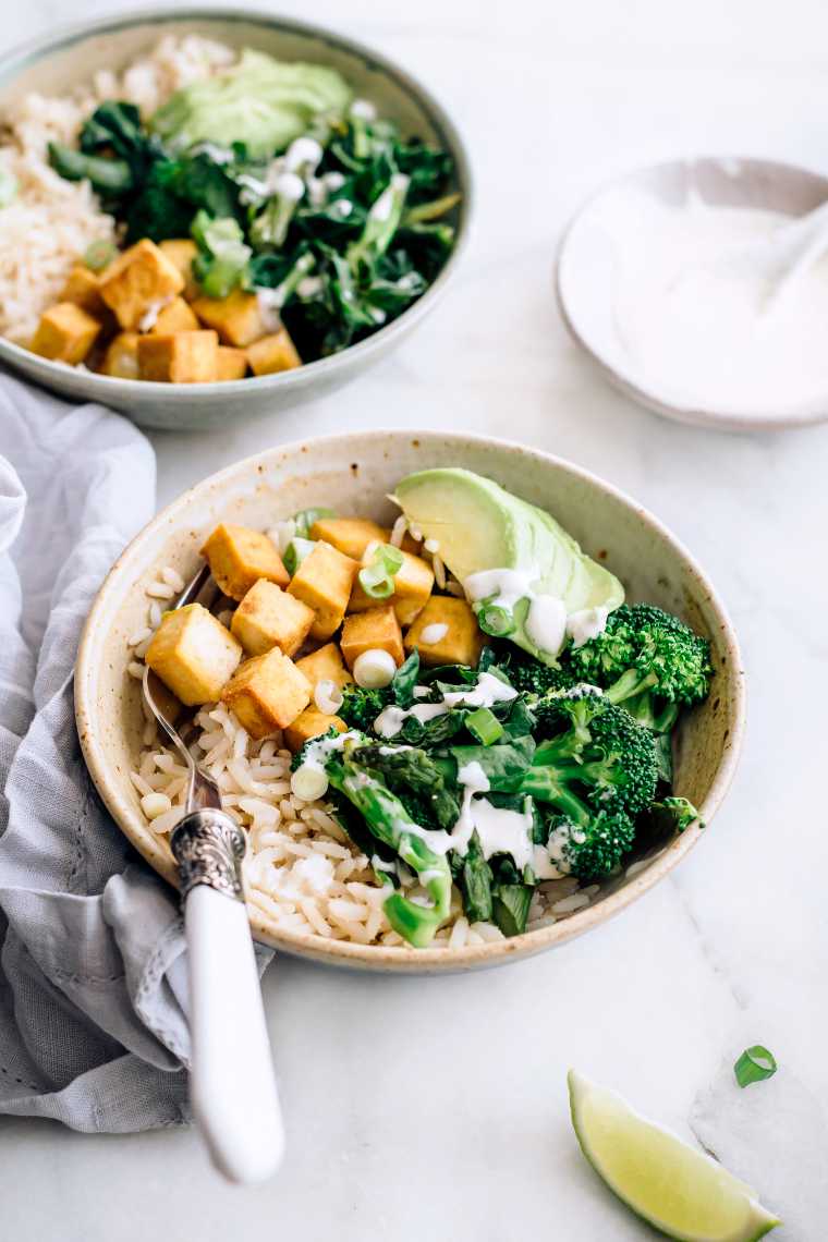 two bowls on a table with rooked rice, green vegetables, sliced avocado and baked tofu, all of which is drizzled with a creamy white vegan sauce