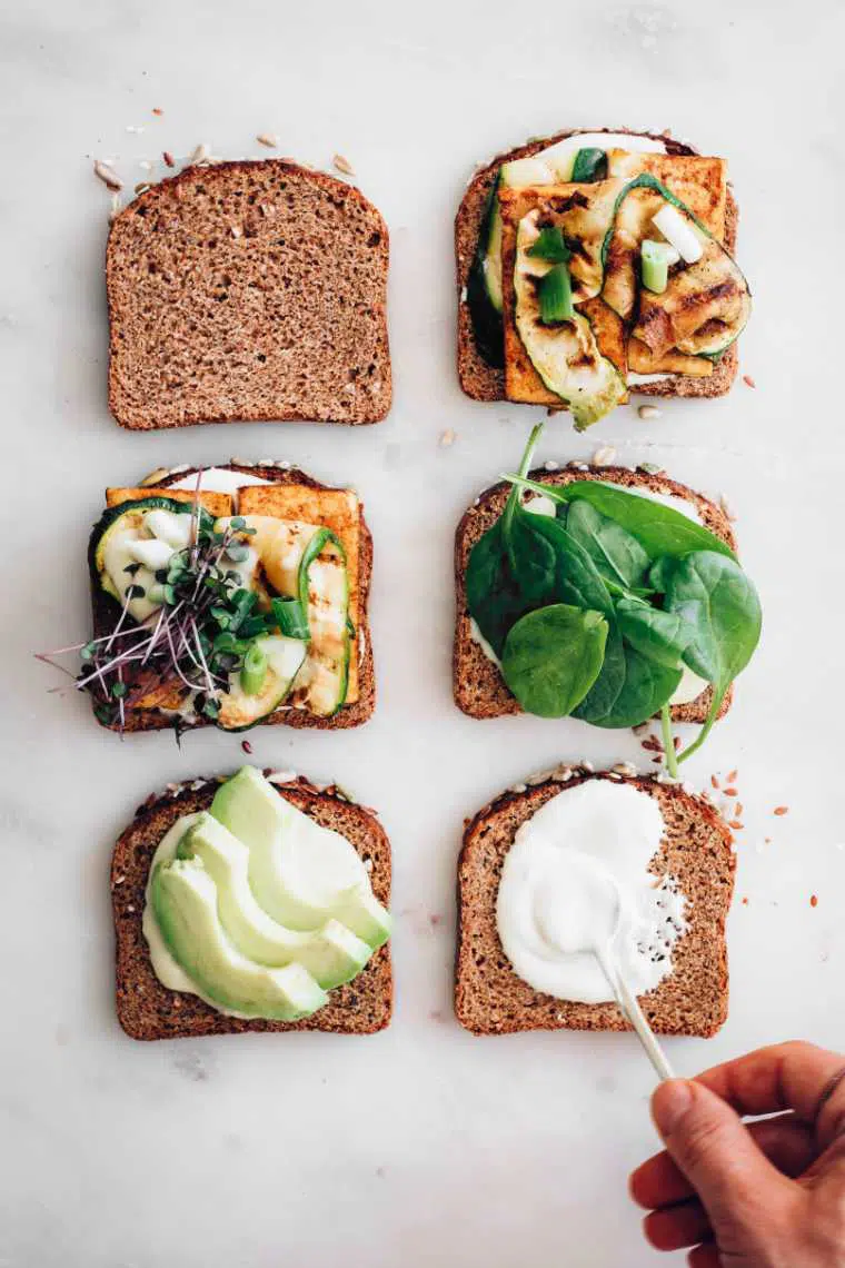 six slices of bread on a white table which are layered with vegan mayo, spinach, avocado, tofu and zucchini