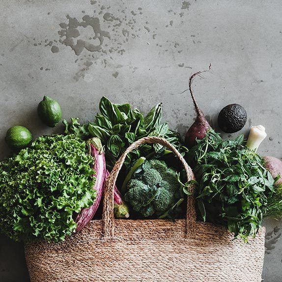 textile grocery bad laying on a wooden surface with fresh produce such as leafy greens, herbs, avocado, broccoli and aubergine
