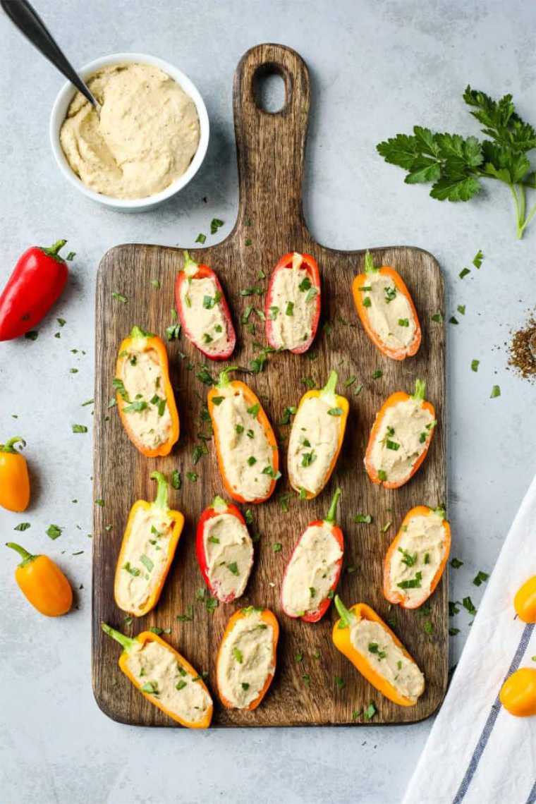 wooden chopping board on grey table with colorful halved mini peppers that are stuffed with hummus